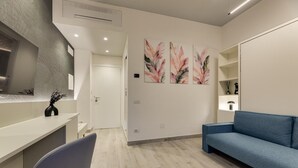 Un salon moderne avec un canapé bleu, un fauteuil gris et une table basse blanche. Trois tableaux aux motifs floraux ornent le mur, et une porte blanche se trouve au fond.