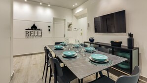 Un espace repas moderne avec une longue table dressée pour quatre personnes, un téléviseur à écran plat fixé au mur et un comptoir de cuisine équipé d’une machine à café.