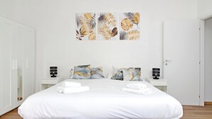 A modern hotel room with a large bed, white bedding, two bedside tables with lamps, and a wall-mounted artwork of tropical leaves.