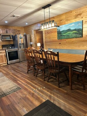 A wooden interior with a dining table and chairs, a kitchen with appliances, and a painting on the wall.