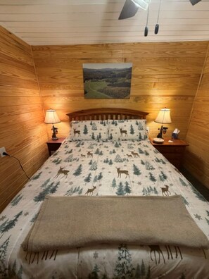 A wooden-paneled room with a bed featuring a deer and tree pattern, two bedside lamps, a painting on the wall, and a headboard.
