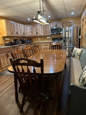 A wooden dining table surrounded by chairs, a kitchen area with cabinets and appliances, and a sofa with cushions.
