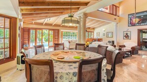 Un vaste espace de vie doté d’un haut plafond, de poutres apparentes, d’une table à manger ronde avec des chaises, et d’une grande fenêtre donnant sur un jardin.