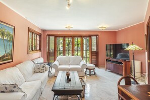 Ein Wohnzimmer mit einem weißen Sofa, einem Couchtisch, einem Fernsehschrank und großen Fenstern mit Rollläden.