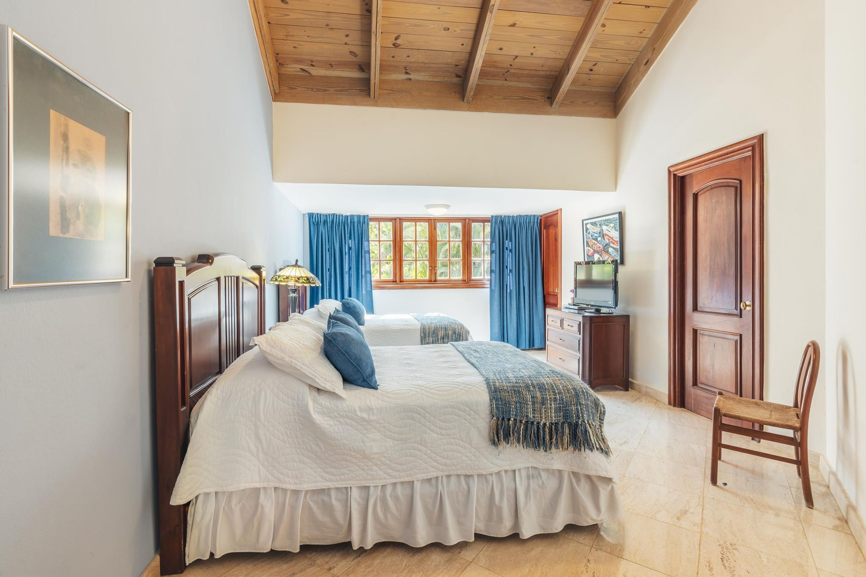 Un dormitorio con una cama grande, techo de madera, ventana con cortinas, televisión y una puerta.
