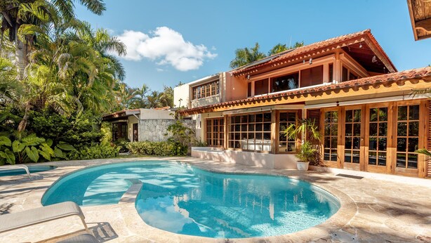 Un espace piscine avec des chaises longues, une maison dotée de grandes fenêtres et une végétation tropicale.