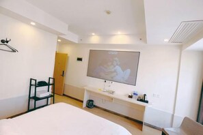 Quarto de hotel com cama, televisão na parede, escrivaninha com cadeira e mesa de cabeceira com abajur.