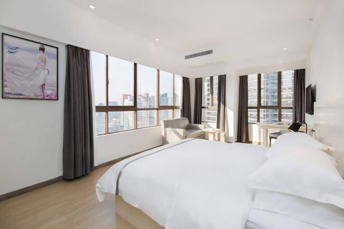 A modern hotel room with a large bed, a desk, a chair, and a view of the cityscape through large windows.