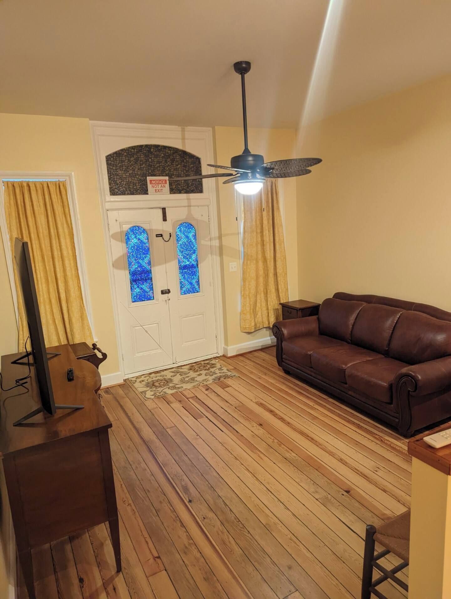 Een woonkamer met een bruine leren bank, een houten vloer, een plafondventilator en een deur met een blauw lichtpatroon.