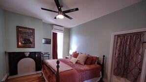 Ein Schlafzimmer mit einem hölzernen Bett, einem Deckenventilator, einem gerahmten Poster und einer Tür mit gemusterten Vorhängen.