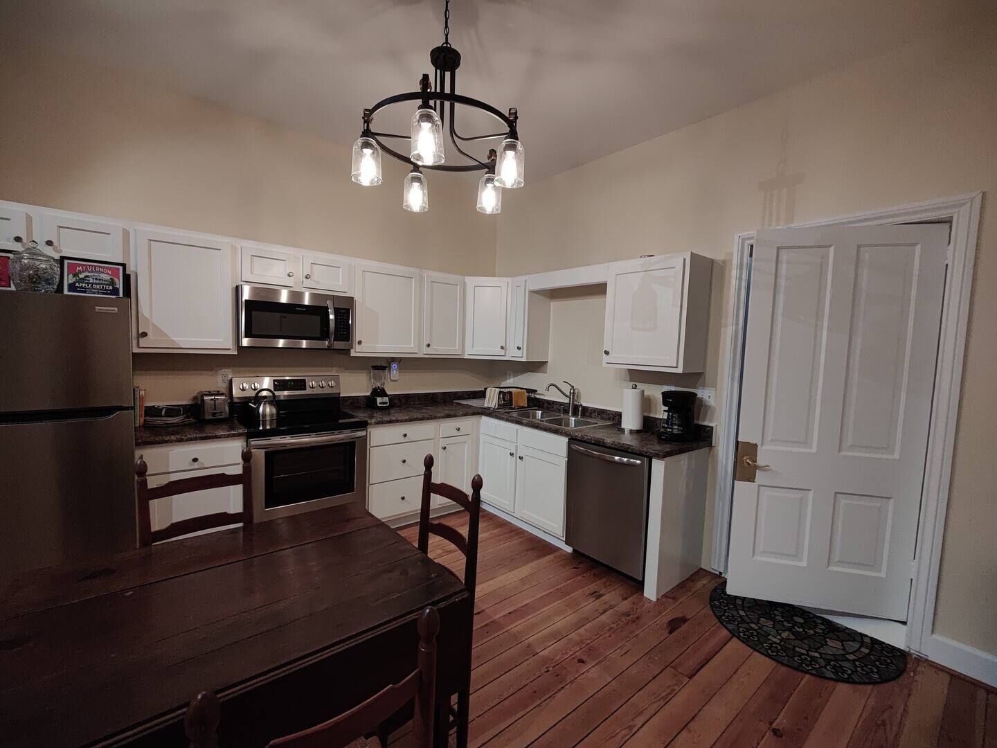 A kitchen with white cabinets, a stainless steel refrigerator, a microwave, a stove, and a dishwasher. There is a wooden dining table with chairs, and a chandelier hanging from the ceiling.