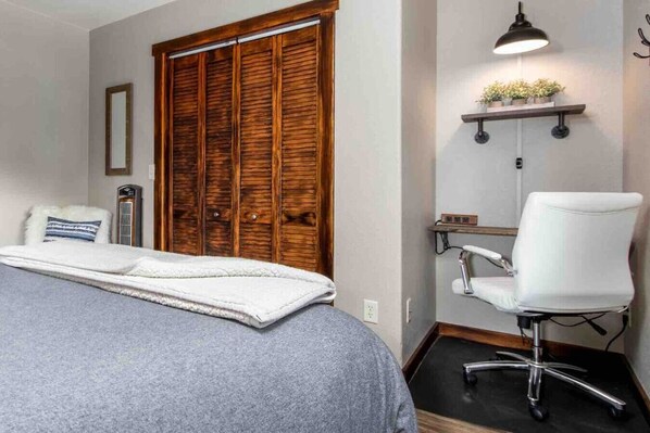 Un dormitorio con una cama, un armario de madera, un escritorio con silla, una estantería en la pared con plantas y un calefactor junto a la cama.