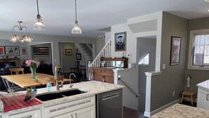 A kitchen with white cabinets, a marble countertop, and a stainless steel dishwasher. There is a wooden dining table with chairs, a vase with flowers, and a hanging light fixture. - 5-Bedroom Estate – 12 acres, Huge Space, Fast WiFi, Fitness Room, & More! (Williamston)