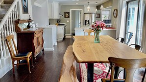 A spacious kitchen with white cabinets, a wooden dining table, and a bench. There is a staircase with white railings on the left, and a vase of flowers on the table. - 5-Bedroom Estate – 12 acres, Huge Space, Fast WiFi, Fitness Room, & More! (Williamston)