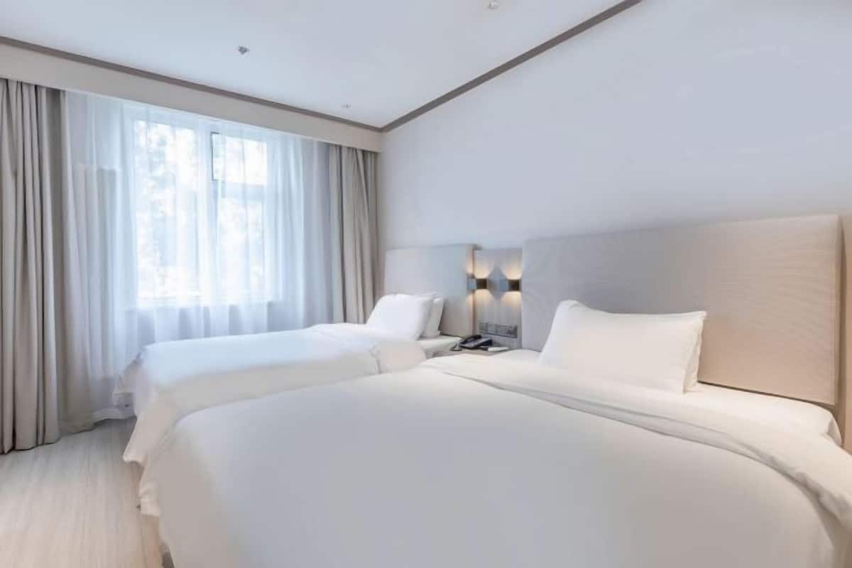 A hotel room with two beds, a large window with curtains, and a wall-mounted lamp.
