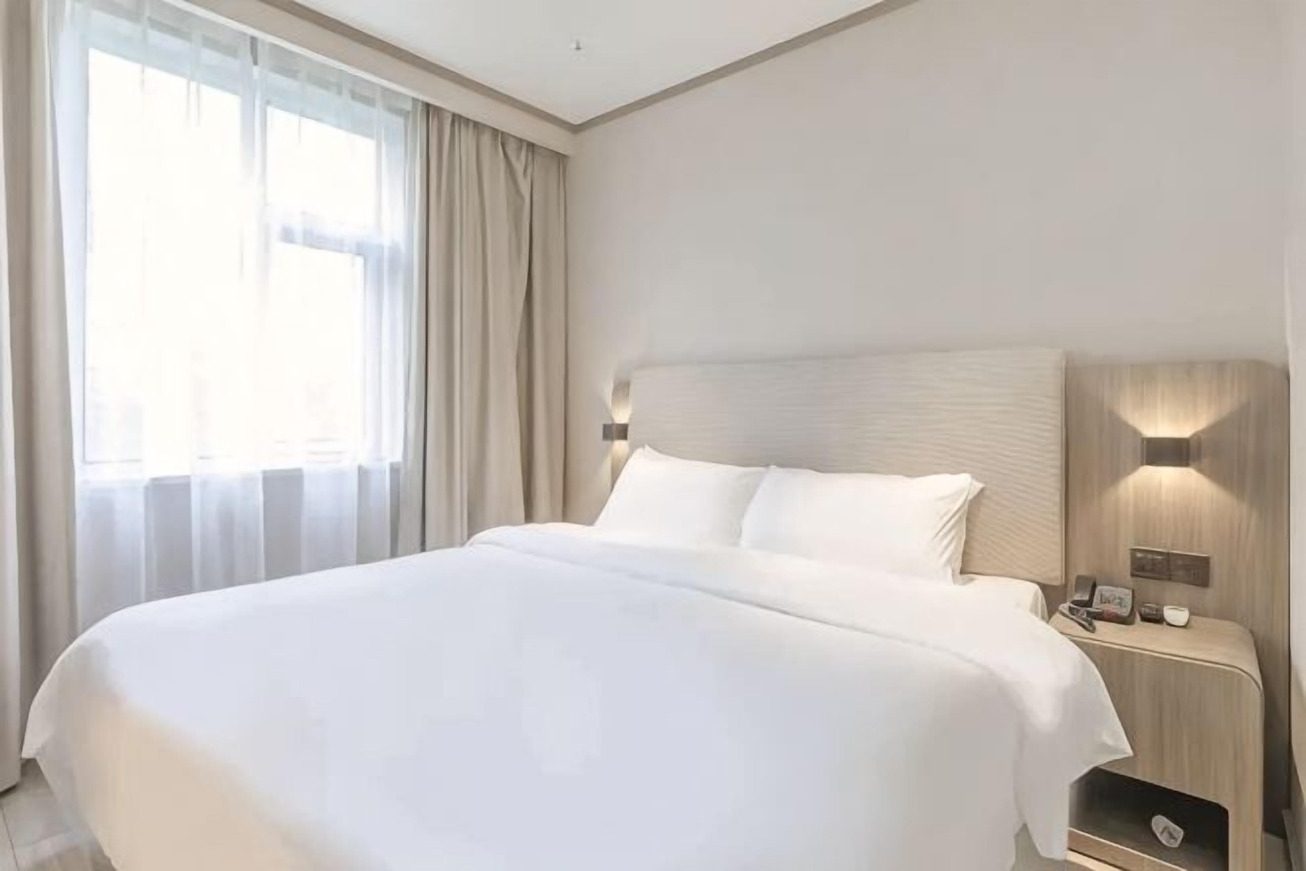 A large bed with a headboard, a bedside table with a lamp, and a window with curtains.