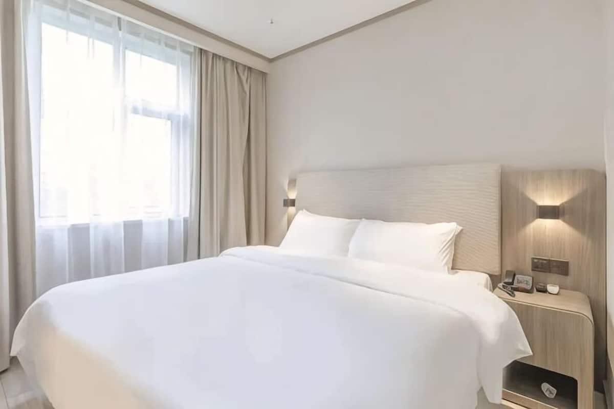 A large bed with a headboard, a bedside table with a lamp, and a window with curtains.