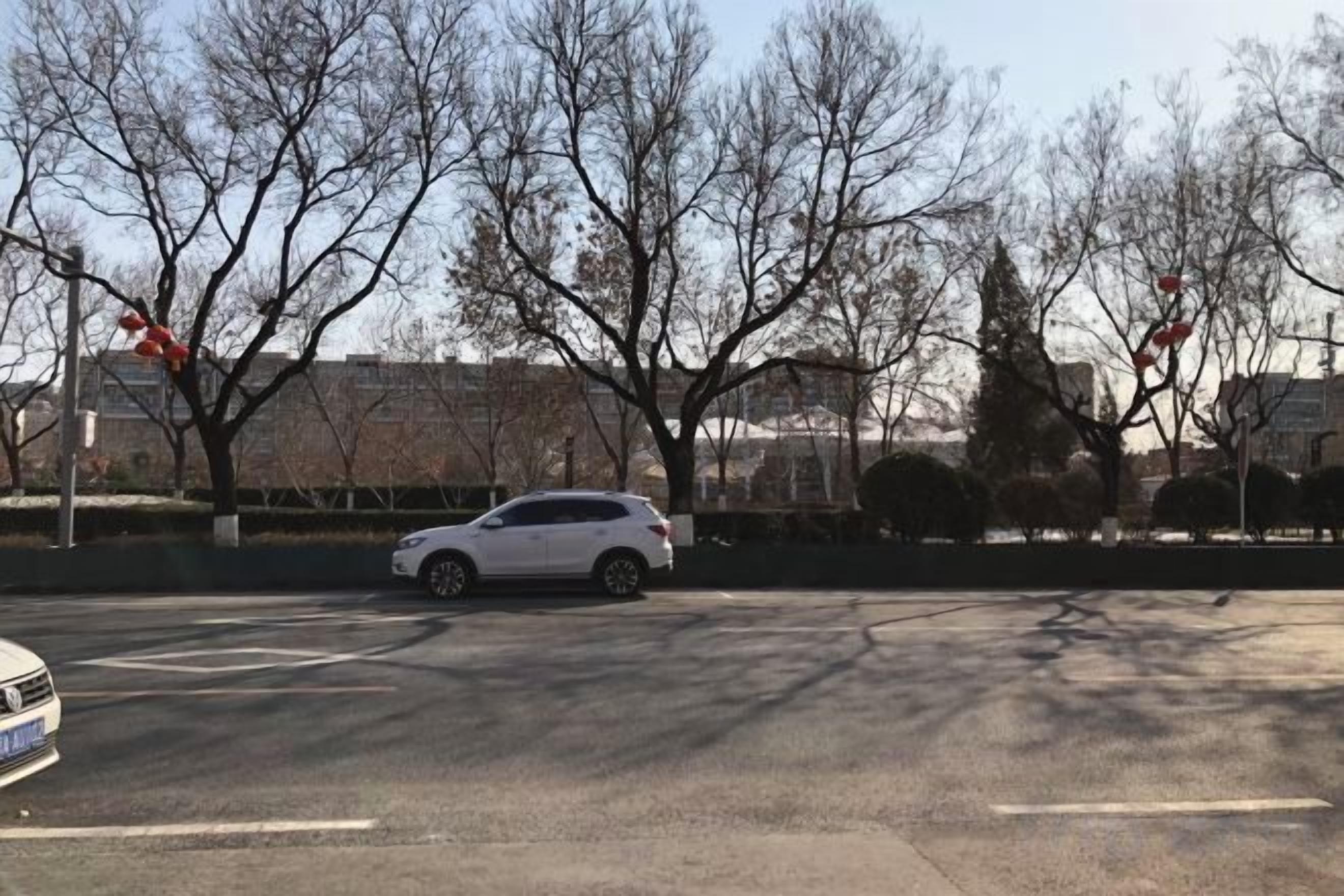 A parking lot with a white car parked near a road intersection, surrounded by leafless trees and a building in the background.