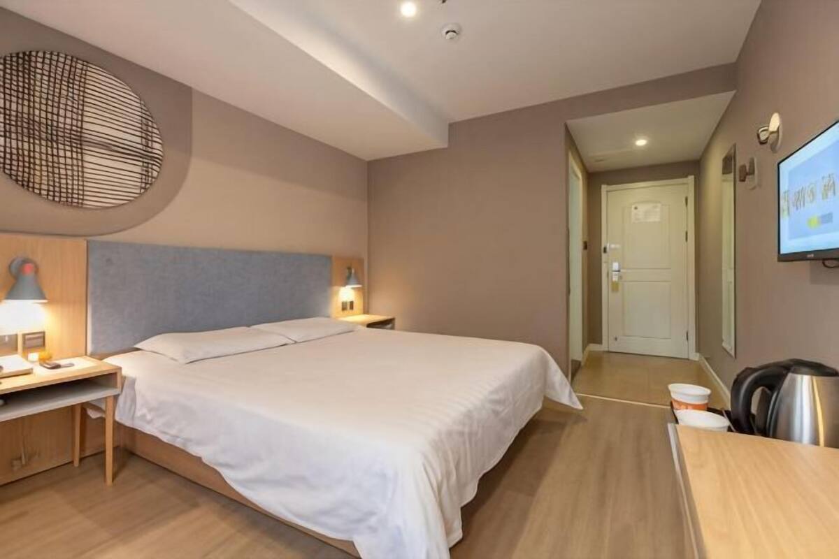 A hotel room with a large bed, bedside tables with lamps, a desk, a chair, a TV, and a door leading to another room.