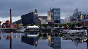 Un paysage urbain au bord de l’eau, avec un mélange de bâtiments historiques et modernes, une rivière paisible qui reflète les structures environnantes et un beffroi imposant.
