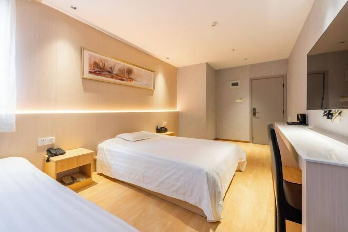 A hotel room with two beds, a bedside table with a phone, a small shelf, a painting on the wall, and a desk with a chair.