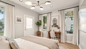 Une chambre avec un grand lit, un bureau et une chaise. Il y a des rideaux aux fenêtres et une plante est posée sur le bureau.