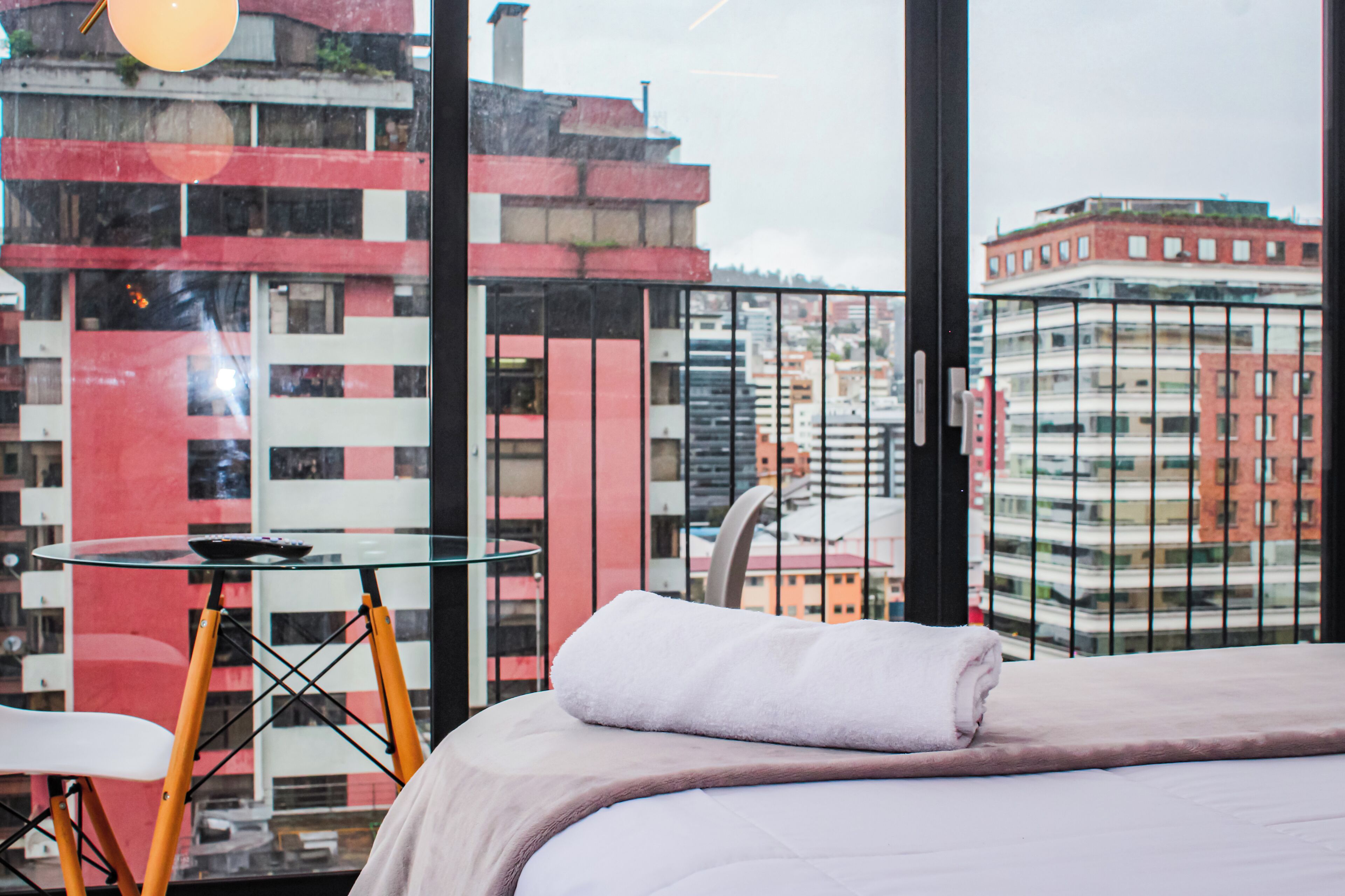 Een hotelkamer met een bed, een glazen tafel en een uitzicht op een stadsbeeld door grote ramen.