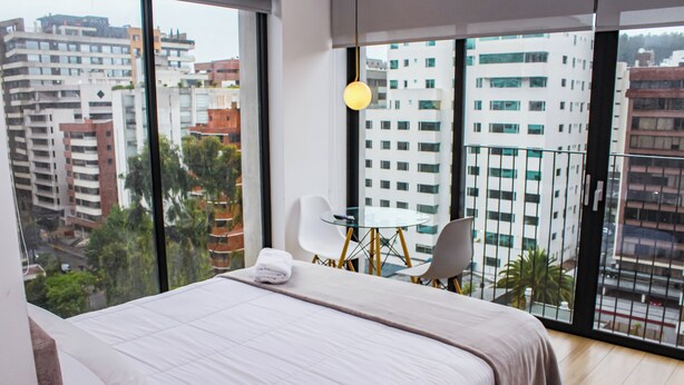Una habitación de hotel moderna con una cama grande, una mesa de vidrio y una silla. La habitación tiene ventanas amplias que ofrecen vistas a la ciudad.