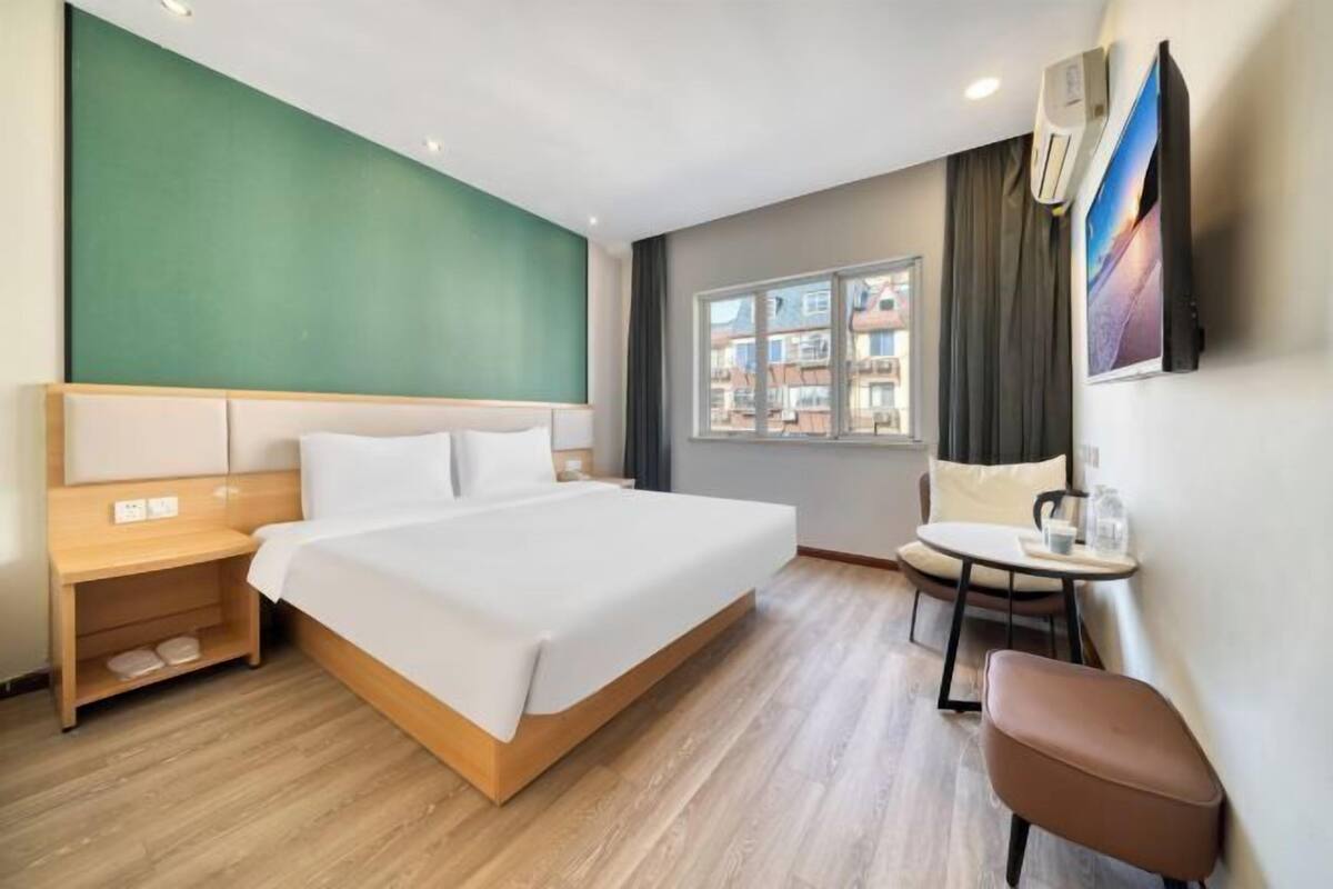A hotel room with a large bed, a small table, a chair, a window with a view of buildings, and an air conditioning unit on the wall.