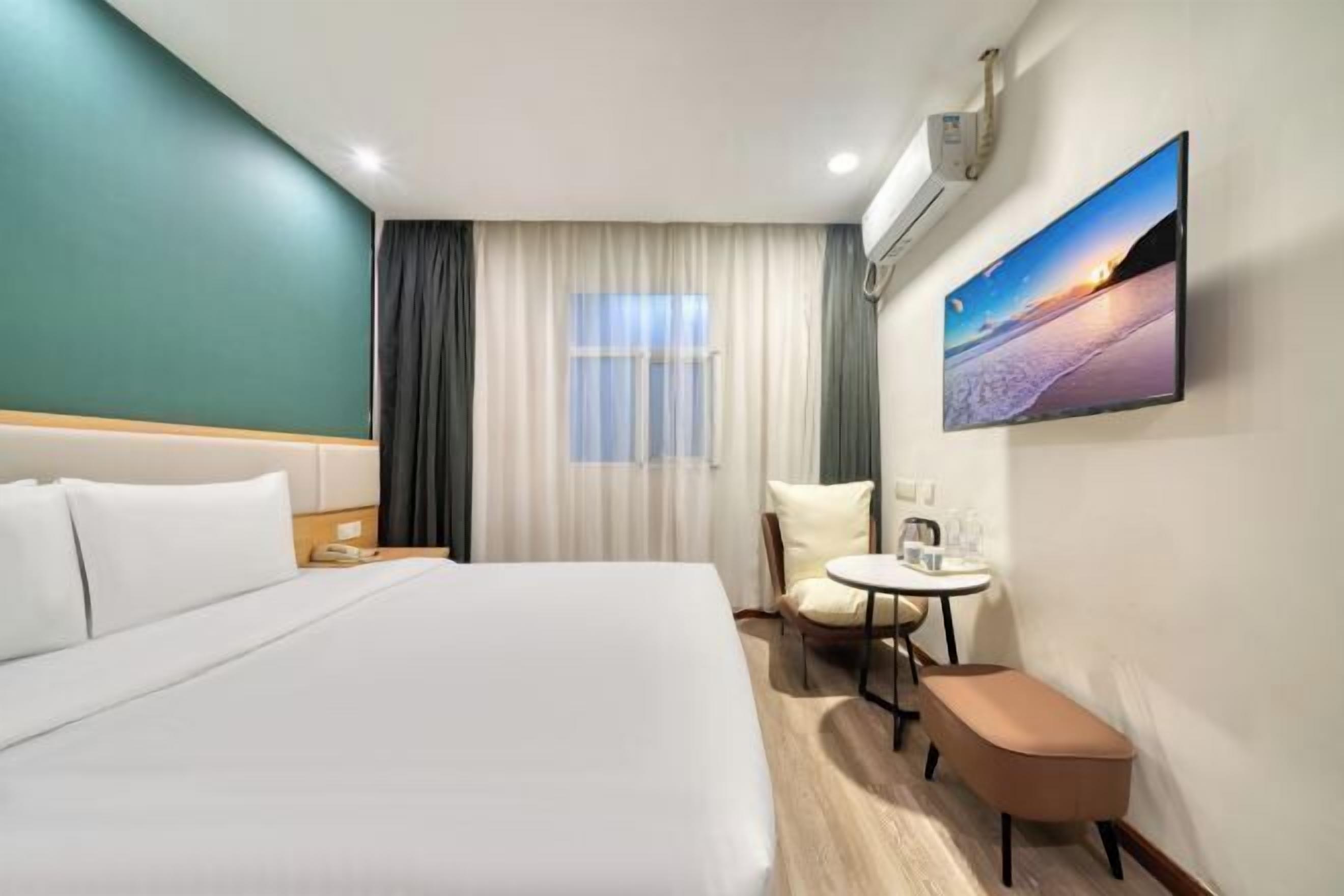 A hotel room with a bed, a small table with chairs, a bench, a window with curtains, and a wall-mounted TV.