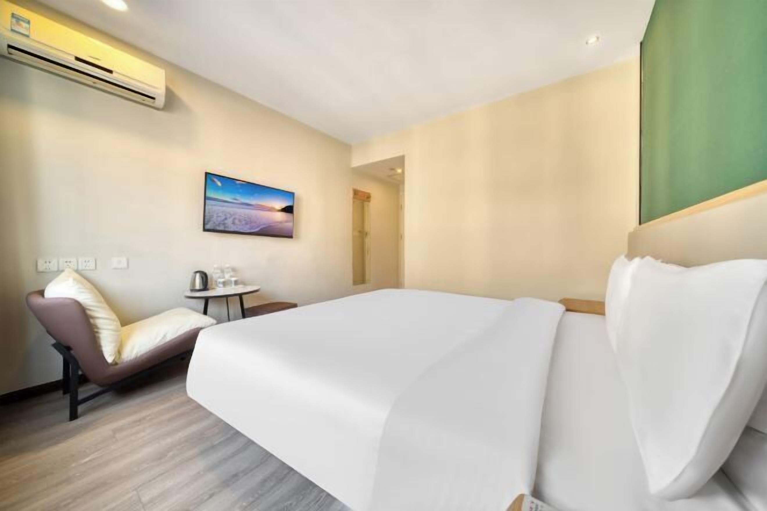 A hotel room with a bed, a chair, a small table with a kettle, and a painting on the wall.