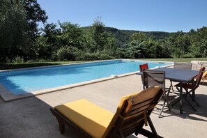 Une piscine rectangulaire à l’eau d’un bleu limpide, entourée d’une terrasse en béton. On y trouve plusieurs chaises longues et une table avec des chaises, dont certaines sont dotées de coussins. La terrasse est bordée d’un petit muret en pierre, et en arrière-plan, on aperçoit de nombreux arbres et des collines verdoyantes.