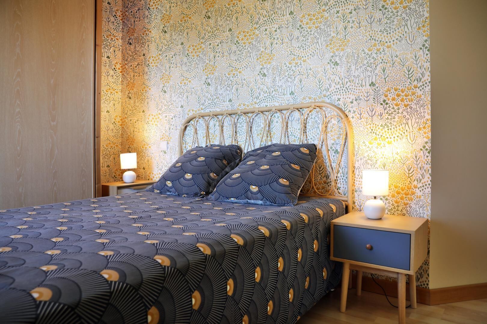 Una camera da letto con un letto, due cuscini, un comodino, una lampada e una parete con motivo floreale.