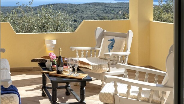 Un balcón con una mesa dispuesta para dos personas, que incluye una botella de vino, copas y un objeto decorativo. El balcón da a un paisaje montañoso.
