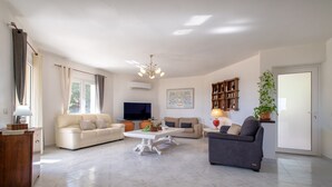 Un salón espacioso con un sofá blanco, un sillón gris, una mesa de centro y un televisor. Se observa una estantería y una planta en maceta.