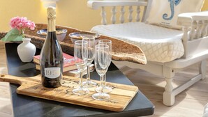 Una bandeja de madera con una botella de vino y copas sobre una mesa, una silla con cojín, y un florero con flores.