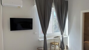 Une pièce équipée d’un téléviseur à écran plat fixé au mur, d’une fenêtre avec des rideaux, d’une chaise en bois et d’un radiateur blanc.