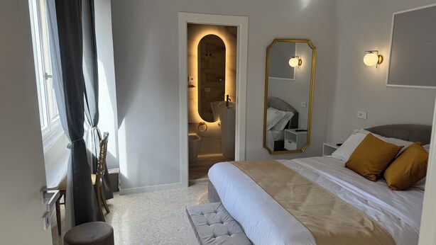 Une chambre d’hôtel avec un lit, un grand miroir, une salle de bain visible à travers une arche et une fenêtre avec des rideaux.