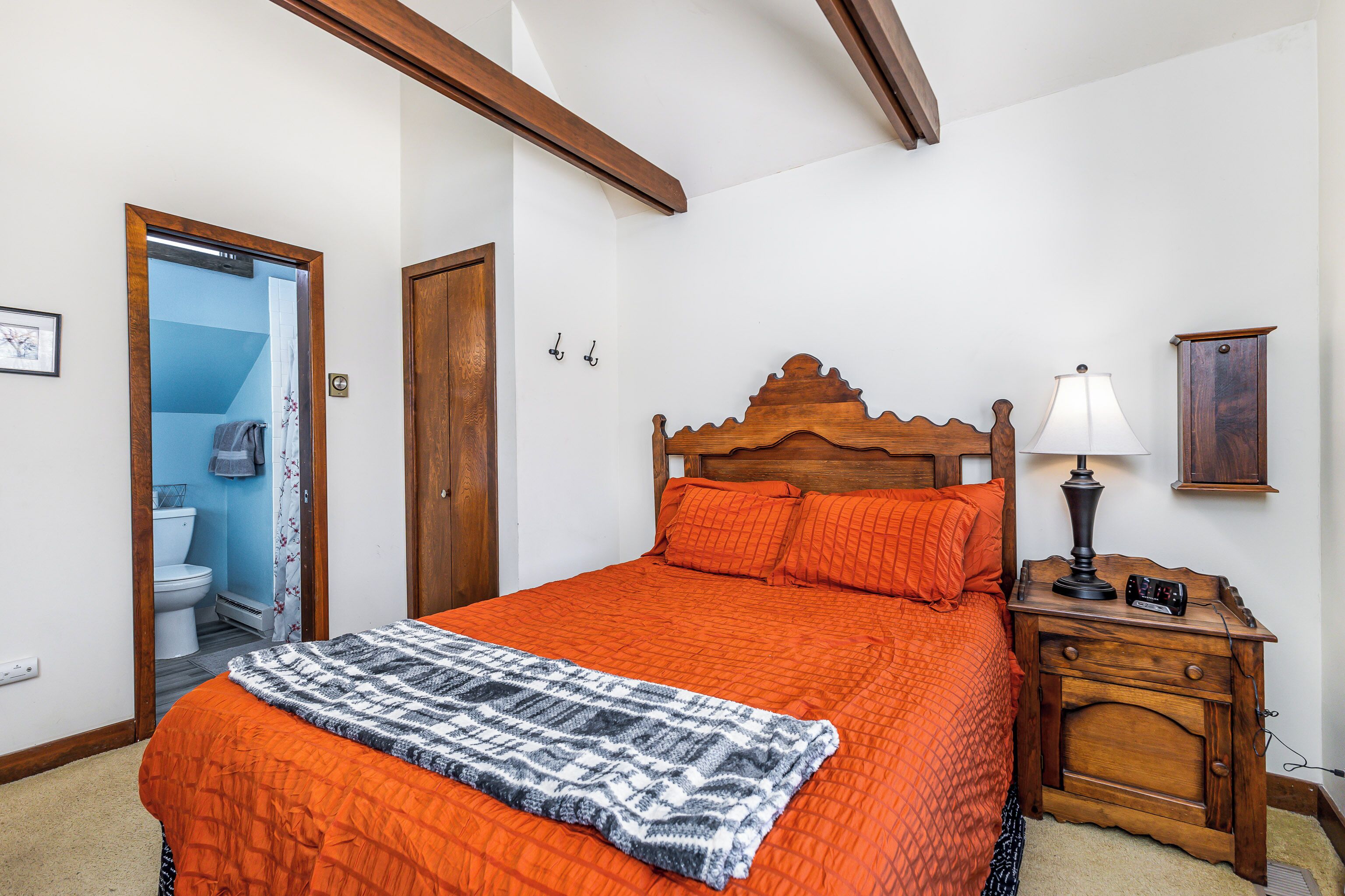 A bedroom with a wooden headboard, an orange bedspread, a patterned throw blanket, a bedside table with a lamp, and a wooden nightstand.