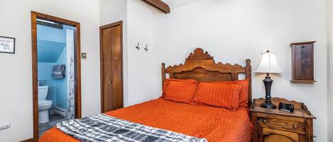 Une chambre à coucher avec une tête de lit en bois, une couvre-lit orange, un plaid à motifs, une table de chevet avec une lampe et une commode en bois.