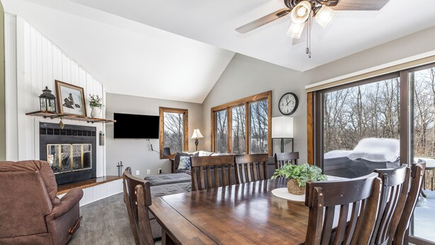 A spacious living room with a wooden dining table, comfortable seating, a fireplace, a television, and large windows offering a view of the outdoors. - Cozy Retreat at Farmstead (Galena)