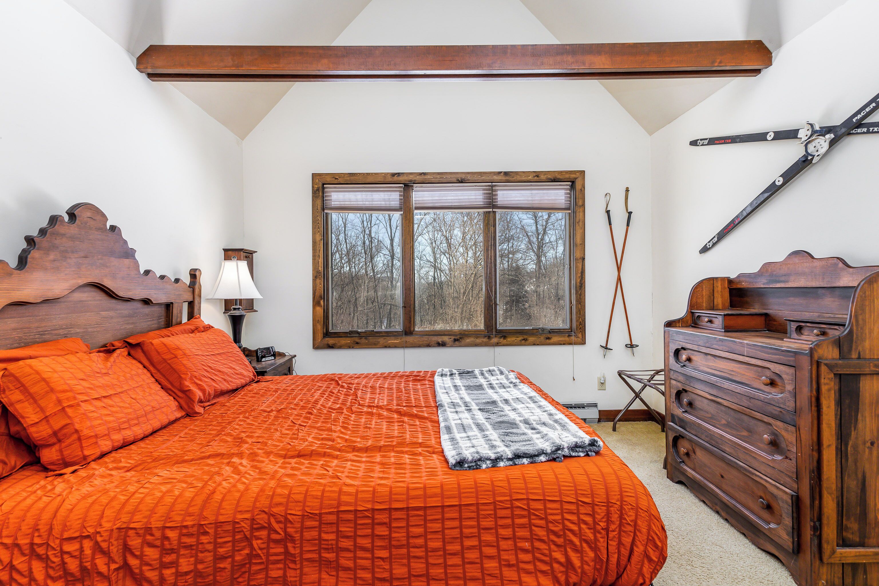 Ein Schlafzimmer mit einem großen Bett, einem Holz-Kopfteil, einem Schminktisch und einem Fenster mit Jalousien. An der Wand hängt ein Ski-Stock.