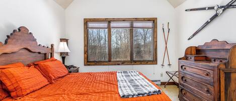 Ein Schlafzimmer mit einem großen Bett, einem Holz-Kopfteil, einem Schminktisch und einem Fenster mit Jalousien. An der Wand hängt ein Ski-Stock.