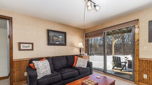A living room with a black sofa, a wooden coffee table, and a large window with a view of a snowy outdoor area. - Cozy Retreat at Farmstead (Galena)