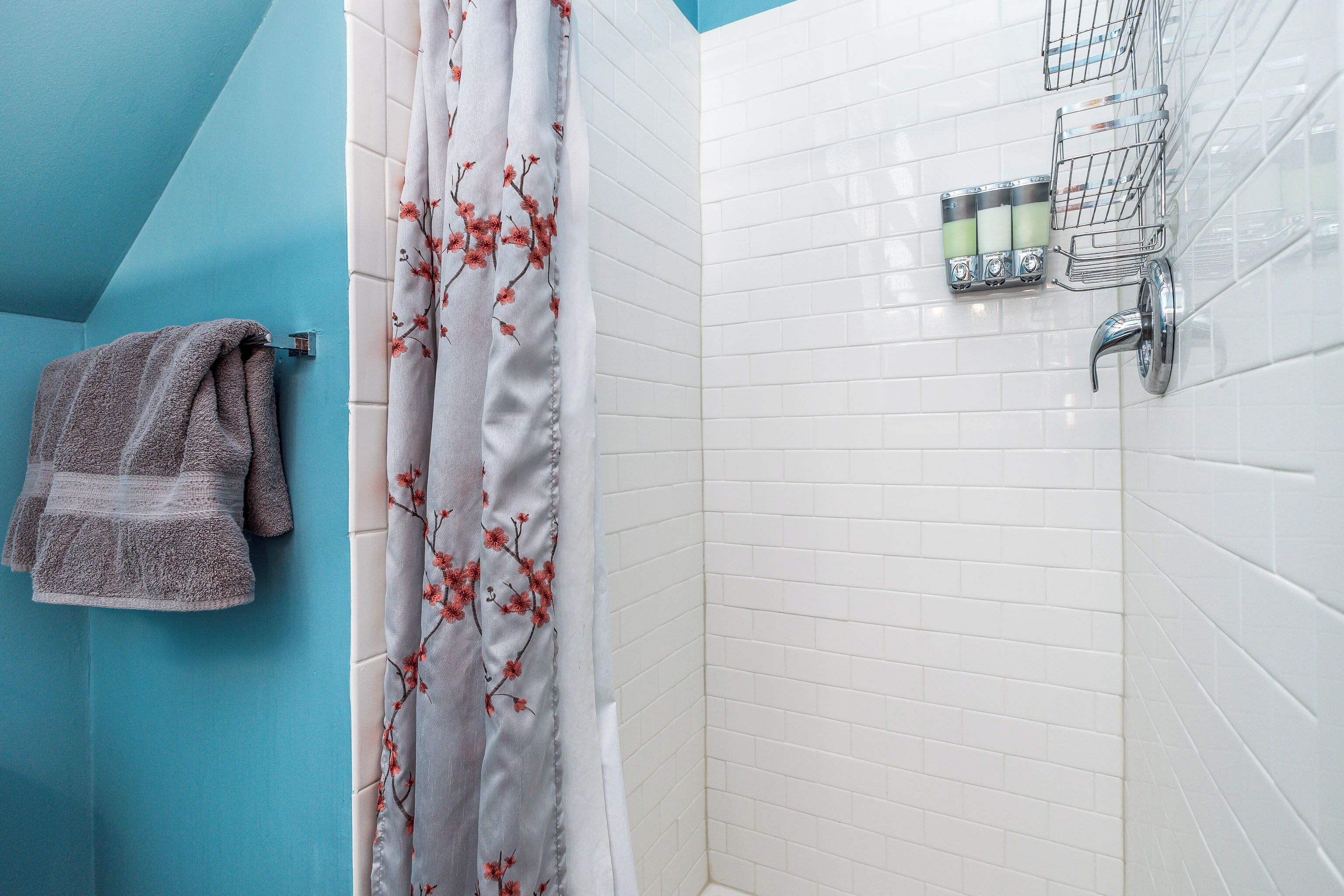 Une salle de bain avec un mur bleu, un espace de douche carrelé de blanc, un rideau de douche à motifs floraux, un porte-serviettes avec une serviette et un distributeur de savon fixé au mur.