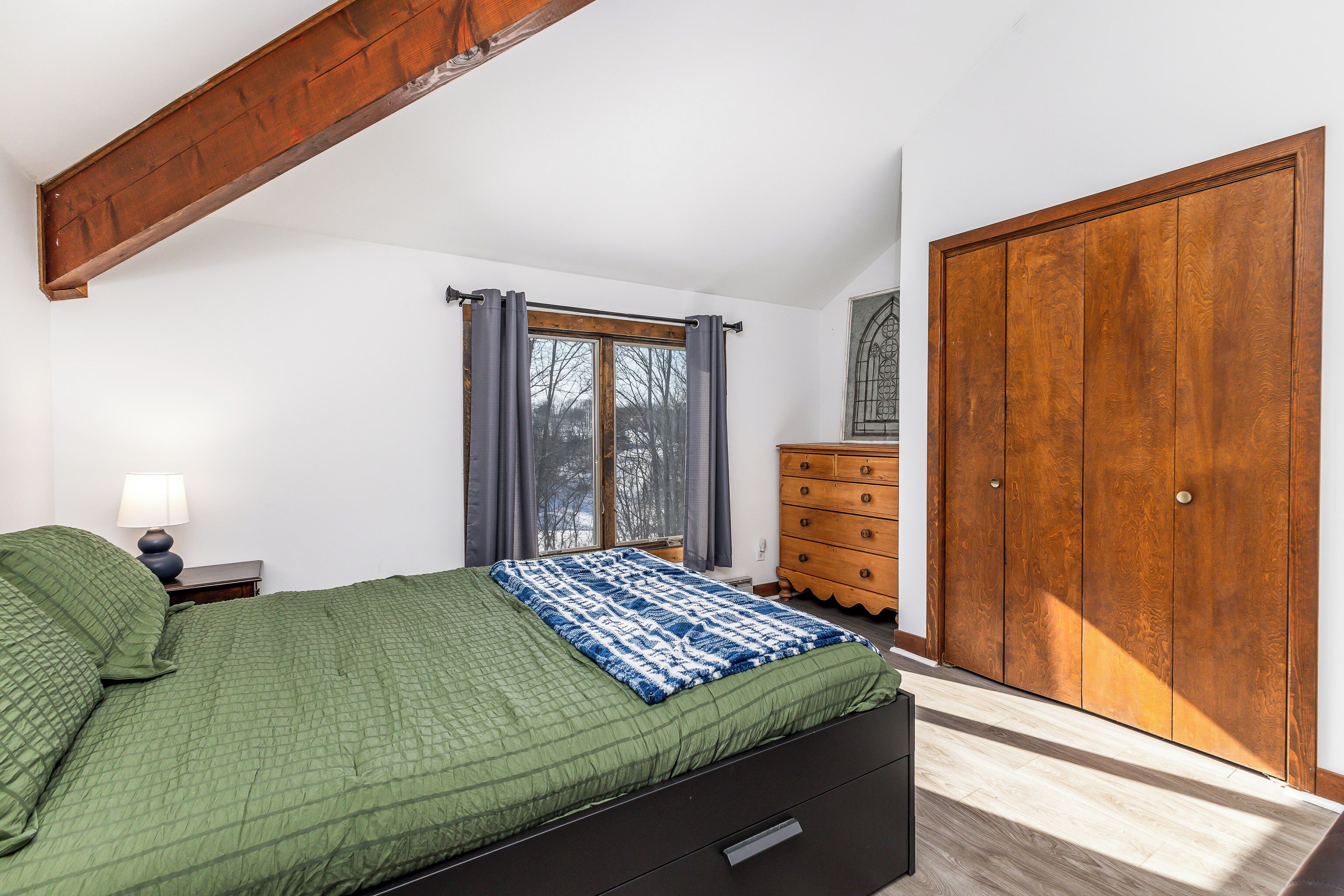 Ein Schlafzimmer mit Bett, Kommode, Kleiderschrank, einem Fenster mit Vorhängen und einer Lampe auf dem Nachttisch.