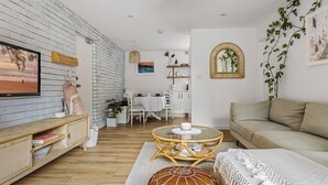 A modern living room with a brick wall, a flat-screen TV, a sofa, a dining area with a white table, and a kitchen area with white cabinets. - The Botanica - Sun-Kissed & Serene (Wollongong)