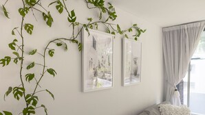A room with a bed, two framed artworks, and a plant with green leaves on the wall. - The Botanica - Sun-Kissed & Serene (Wollongong)