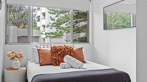 A bedroom with a large window, a bed with white and brown bedding, a vase with flowers, and a mirror on the wall. - The Botanica - Sun-Kissed & Serene (Wollongong)