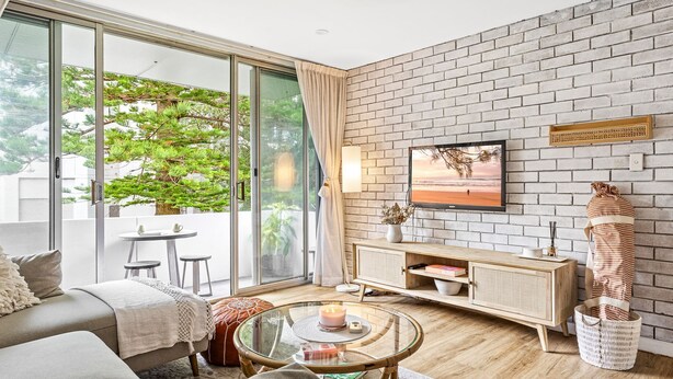 A modern living room with a glass door leading to a balcony, a brick wall, a flat-screen TV, and a cozy seating area. - The Botanica - Sun-Kissed & Serene (Wollongong)